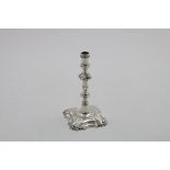 A GEORGE II CAST TAPERSTICK on a shaped square base with shell decoration, a knopped column and a