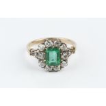 AN EMERALD AND DIAMOND CLUSTER RING, the rectangular step-cut emerald claw set within a border of