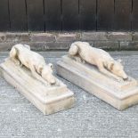 A pair of reconstituted stone garden models of lying greyhounds, each on a plinth base,