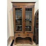 An early 20th Arts & Crafts oak display cabinet enclosed by leaded glass doors over drawer and