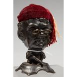 A GERMAN CAST IRON AND MAROON VELVET HATPIN STAND AS A CARICATURE OF A BESPECTACLED, SEATED