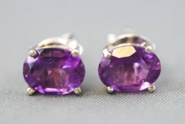 A white metal pair of single stone stud earrings. Each set with an oval faceted cut amethyst.