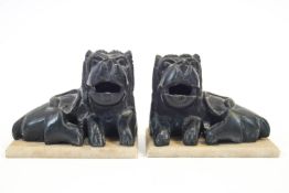A pair of Asian style black stoneware figures of recumbent lions on white marble bases,