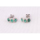 A white metal pair of stud earrings each set with three oval faceted cut emeralds