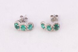 A white metal pair of stud earrings each set with three oval faceted cut emeralds