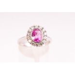 A white metal cluster ring. Set with an oval faceted cut pink sapphire and surrounded by diamonds.