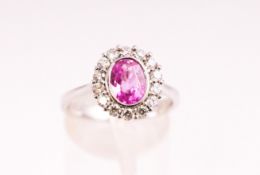 A white metal cluster ring. Set with an oval faceted cut pink sapphire and surrounded by diamonds.