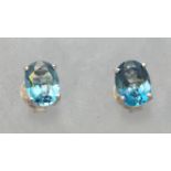 A white metal pair of single stone stud earrings. Each set with an oval faceted cut blue topaz.