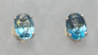 A white metal pair of single stone stud earrings. Each set with an oval faceted cut blue topaz.