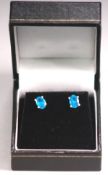 A white metal pair of single stone stud earrings. Each set with an oval cut intense blue apatite.