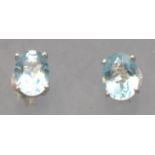 A white metal pair of single stone stud earrings. Each set with an oval faceted cut pale blue topaz.