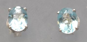 A white metal pair of single stone stud earrings. Each set with an oval faceted cut pale blue topaz.