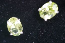 A white metal pair of single stone stud earrings. Each set with an oval faceted cut peridot.