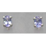 A white metal pair of single stone stud earrings. Each set with an oval cut tanzanite.