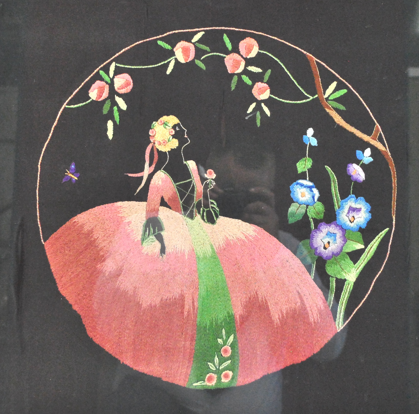 An Art Deco silk work of a lady in a crinoline dress, in a garden setting with roses and hollyhocks,