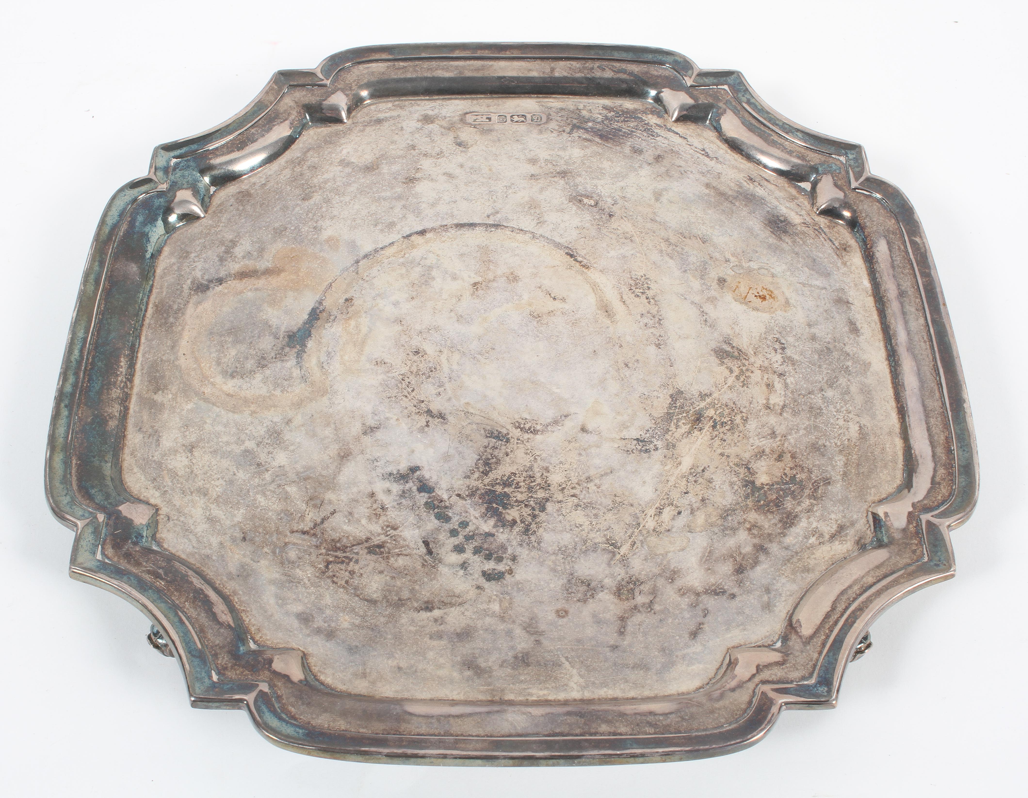A silver salver, of cut cornered square design with fancy border, raised on four scroll feet,
