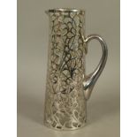 AN EARLY 20TH CENTURY SILVER OVERLAID GLASS WATER JUG, tapered cylindrical outline, the scrollwork