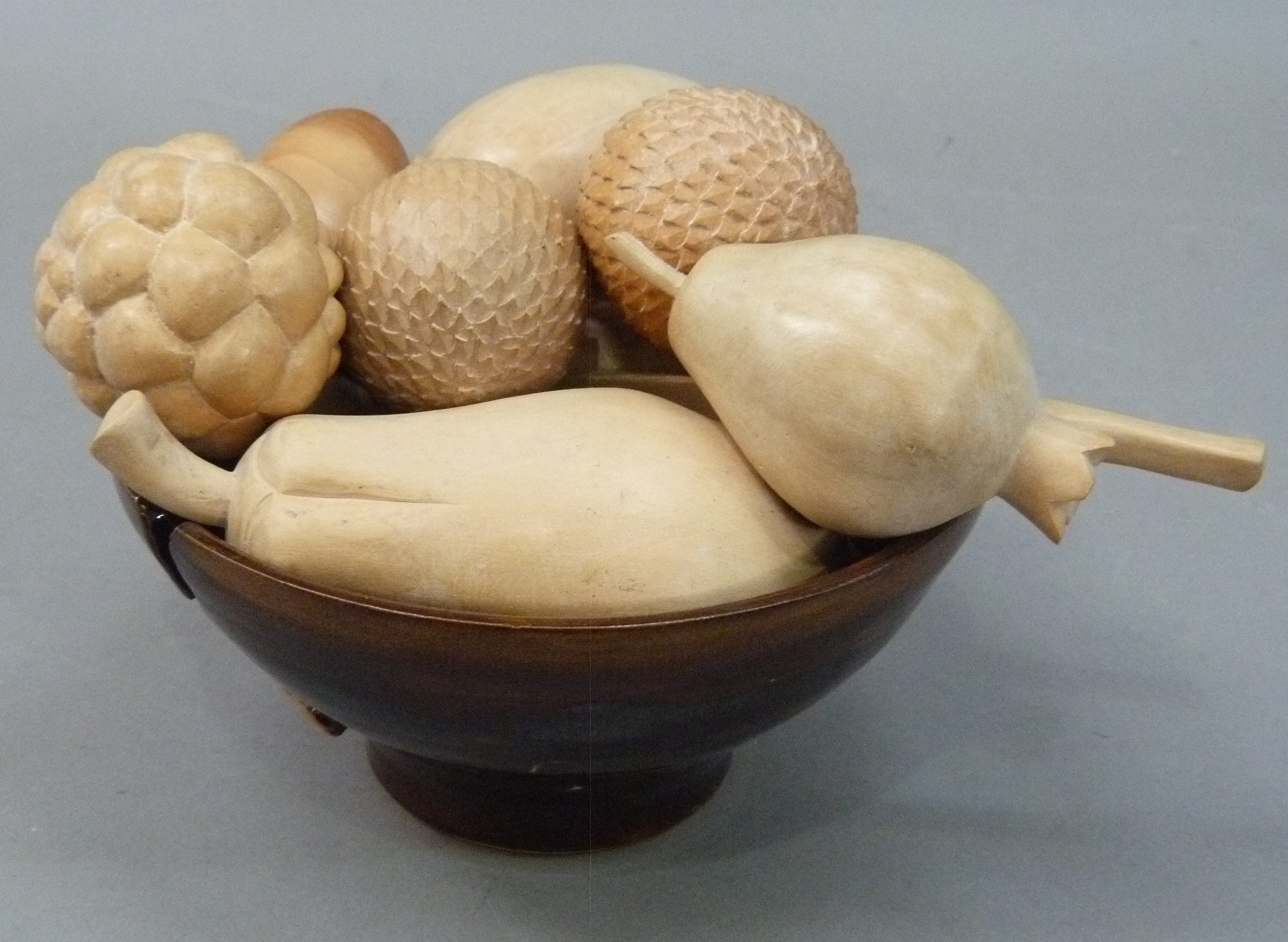 A studio pottery bowl of brown glaze containing a collection of hand carved wooden fruits