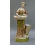 A Royal Dux figure of a young woman standing holding an oval basket, another basket to her left,