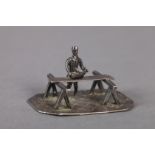 A LATE 19TH CENTURY MINIATURE SILVER FIGURE OF A CARPENTER, planing a length of wood resting on