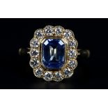 A SAPPHIRE AND DIAMOND CLUSTER RING in 18ct gold, the step cut sapphire collet set within a surround