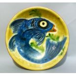 A studio pottery plate decorated with a blue fish on a mustard ground, 27cm diameter
