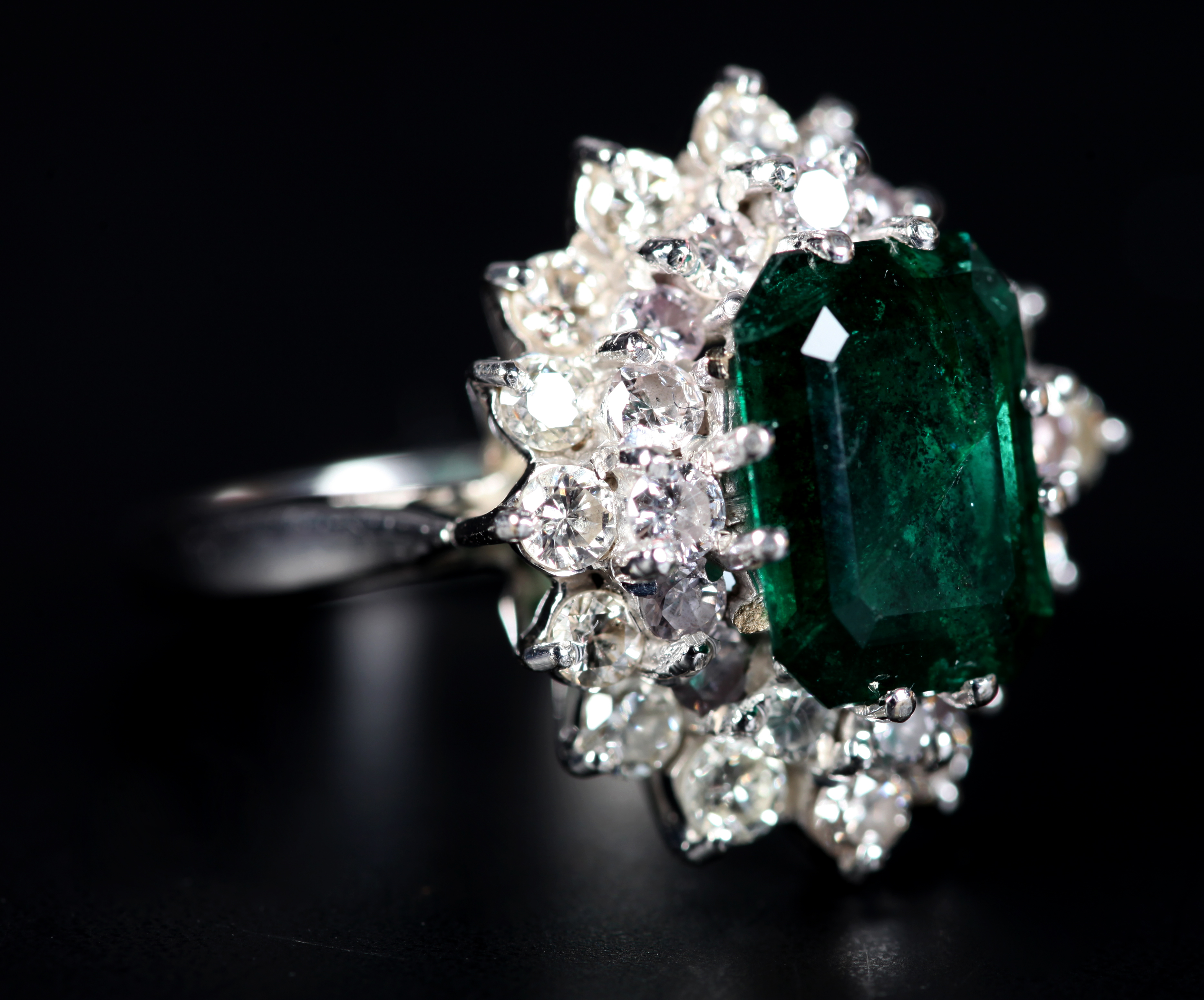 AN EMERALD AND DIAMOND CLUSTER RING claw set to the centre with a step cut diamond within an