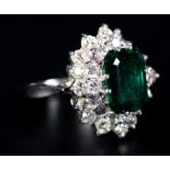 AN EMERALD AND DIAMOND CLUSTER RING claw set to the centre with a step cut diamond within an
