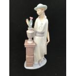 A large Lladro figurine of a lady in a long flowing dress and hat looking at a vase of flowers on