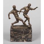 A bronzed metal figure group of a pair of football players raised on a tall rectangular marble