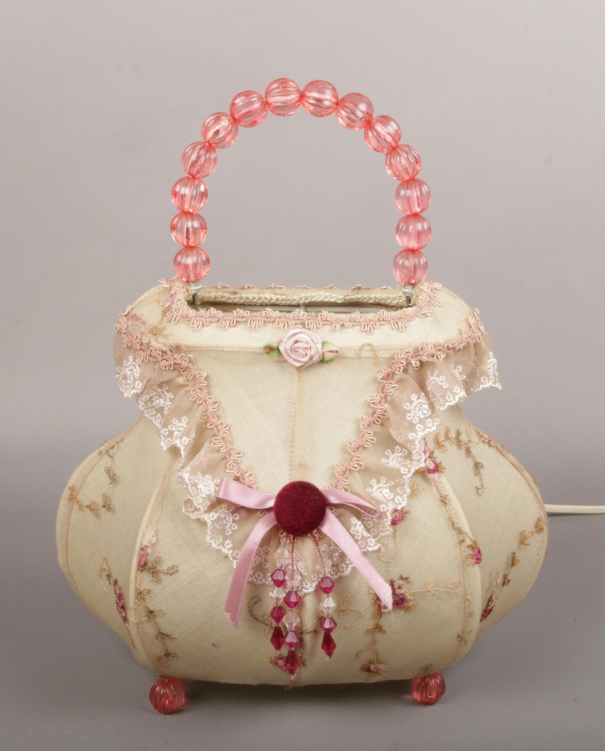 A novelty tablelamp formed as a handbag. With beadwork handle and feet and having embroidered lace