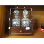 A MAHOGANY VENEERED TANTALUS WITH A PAIR OF GLASS STOPPERED DECANTERS AND SILVER DECANTED LABELS,