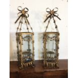 A pair of wrought iron hanging lights, the square glass enclosures surmounted by ribbon bows, the