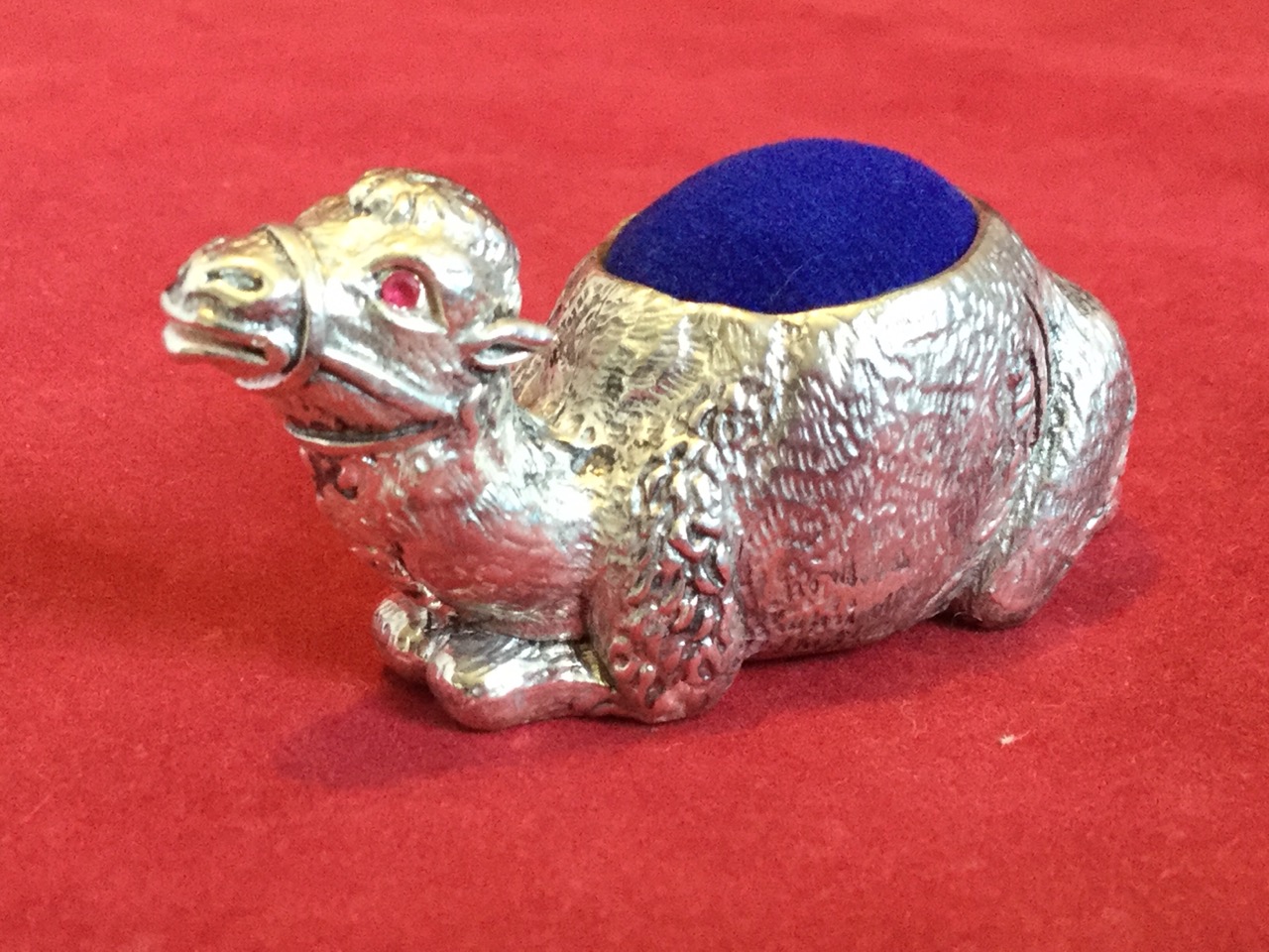 A silver plated pin cushion cast as a seated camel having inlaid ruby eyes, the hump or back with