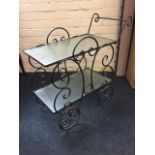 A wrought iron drinks trolley with two rectangular mottled glass shelves in scrolled frame on four