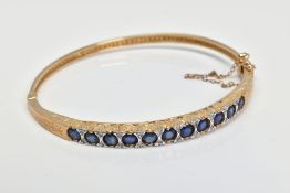 A 9CT GOLD SAPPHIREM AND DIAMOND HINGED BANGLE, designed as a line of eleven circular sapphires