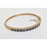 A 9CT GOLD SAPPHIREM AND DIAMOND HINGED BANGLE, designed as a line of eleven circular sapphires