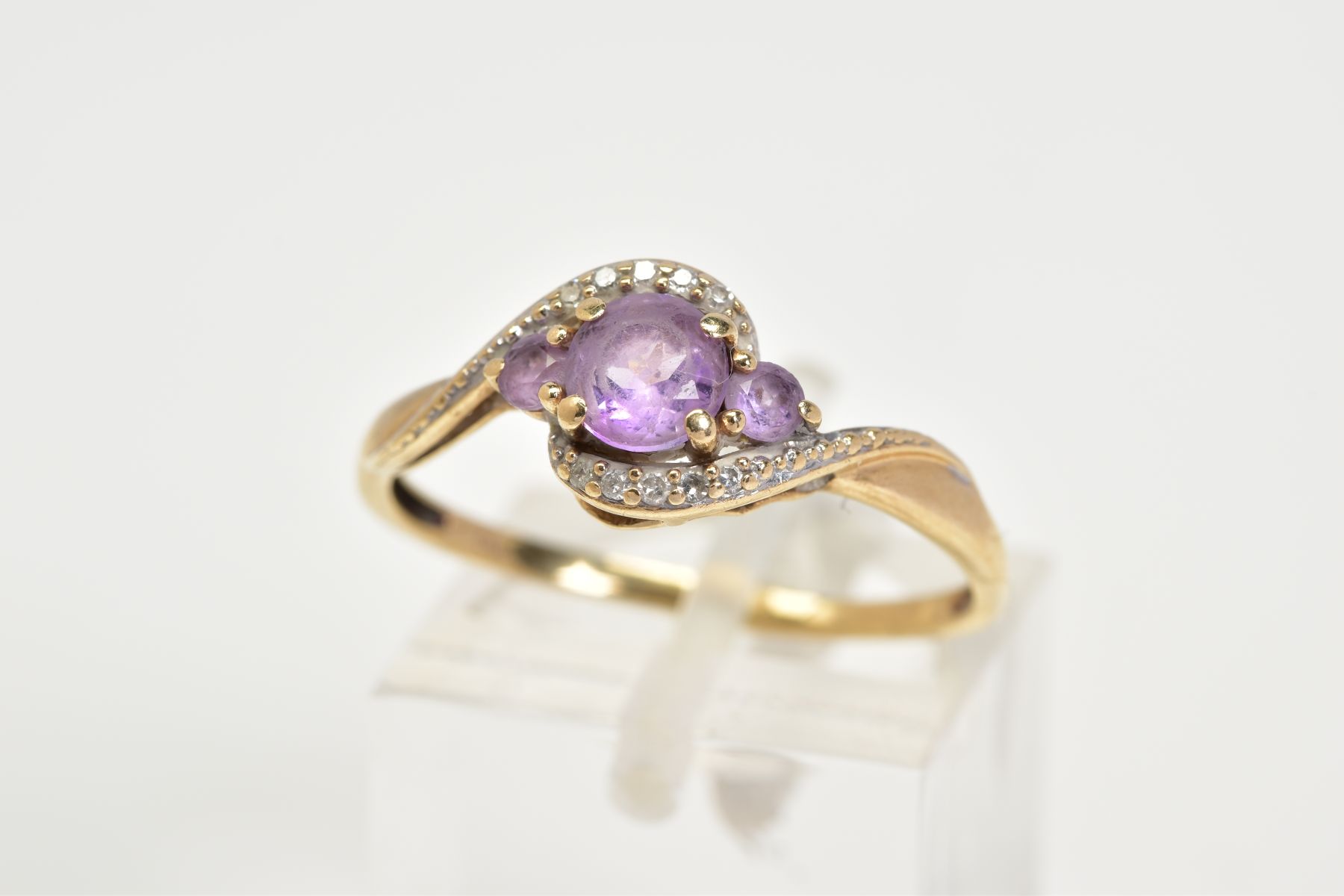 AN AMETHYST AND DIAMOND RING, the yellow metal ring of cross over design, set with three graduated