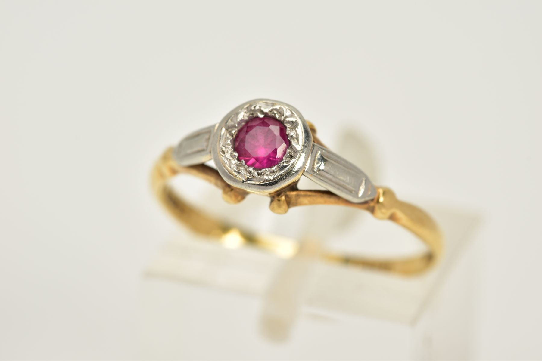 A YELLOW METAL RUBY RING, designed with a single circular cut ruby within a collet mount, textured
