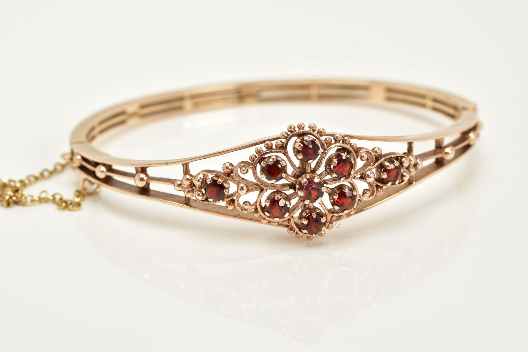 A 9CT GOLD GARNET SET OPENWORK BANGLE, the openwork hinged bangle of floral design, set with