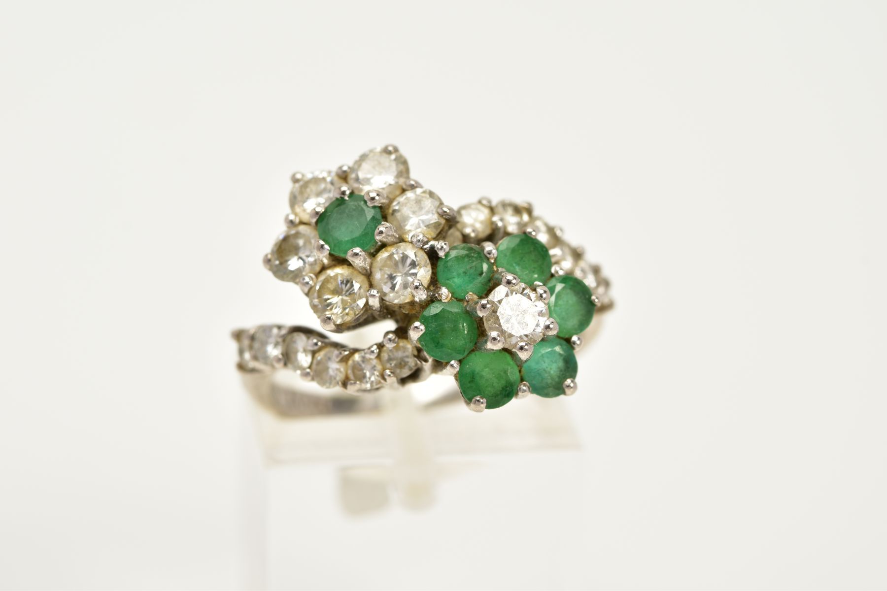 A DIAMOND AND EMERALD CROSSOVER RING, the white metal ring of crossover design, in the form of two