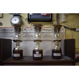A set of three plated metal twin handled pedestal trophy cups, each on wood socle, total height
