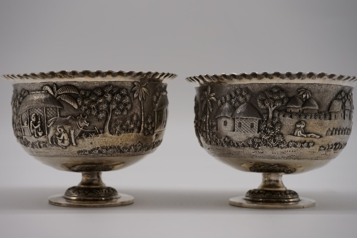 A pair of Indian white metal pedestal bowls, decorated village scenes, 8.5cm high.