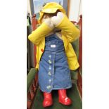 A large stuffed doll in denim dress, pvc mac and hat with red wellies. The doll's arms attached to