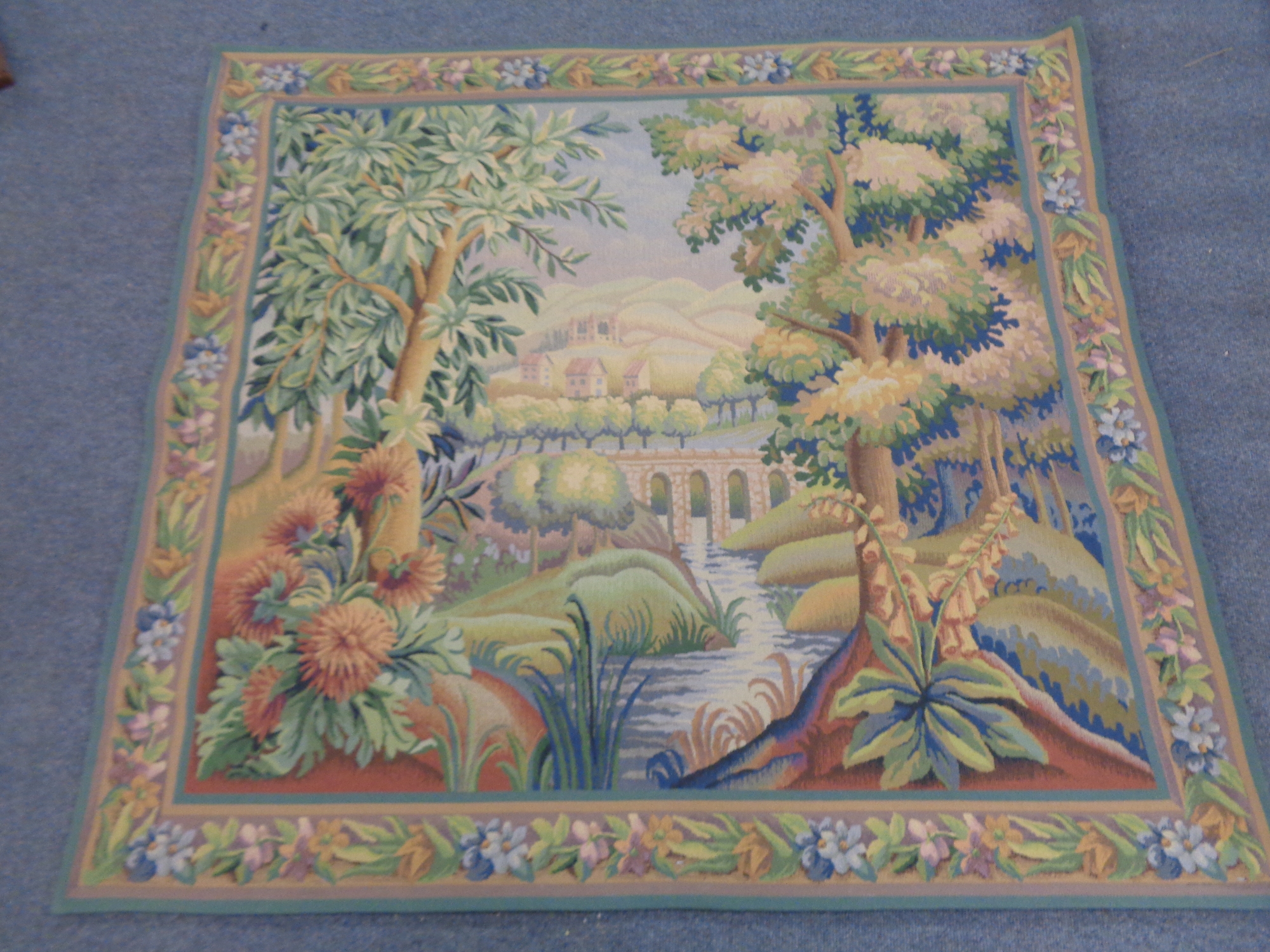 A wall tapestry depicting a river through a woodland with bridge and houses beyond