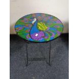 A folding metal occasional table with glass panel top depicting a peacock