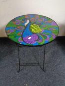 A folding metal occasional table with glass panel top depicting a peacock