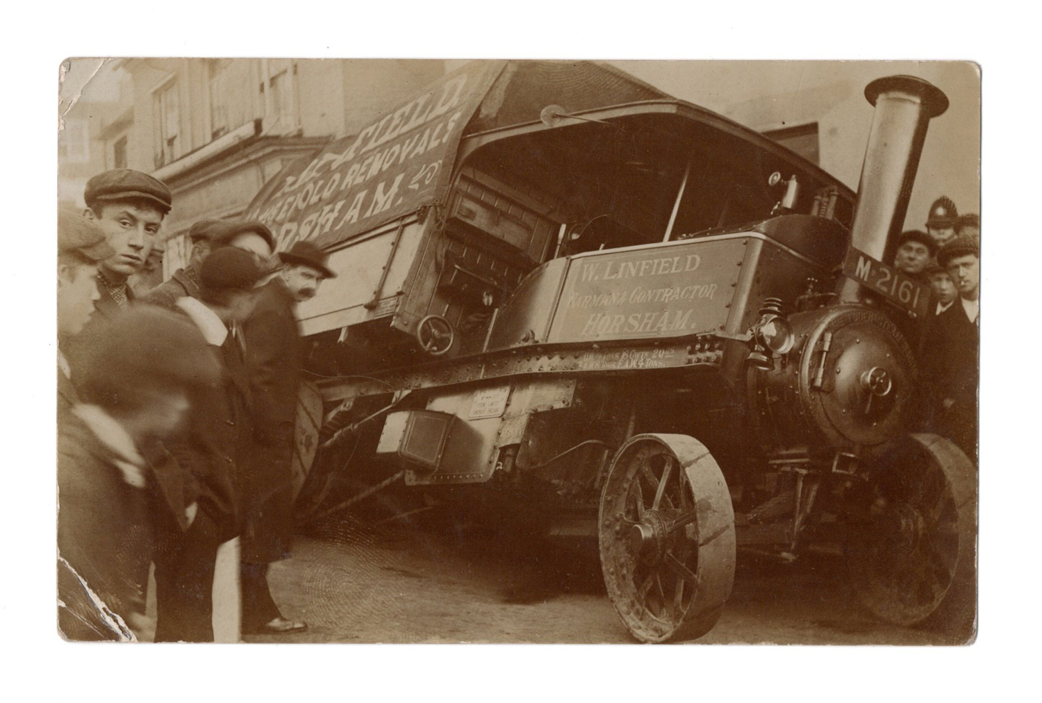 A photographic postcard of the partially overturned W. Linfield Removals steam engine in Horsham.