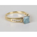 A gold, pale blue gem and diamond ring, claw set with the square step cut blue stone between diamond