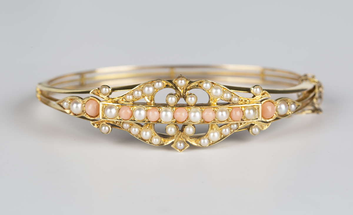 A gold, coral and seed pearl oval hinged bangle, circa 1910, the front in a pierced scrolling